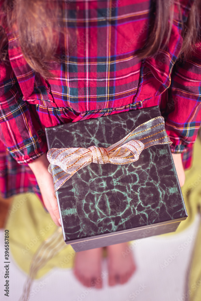 A little girl in a red plaid dress holding a Christmas gift box with ribbon.
