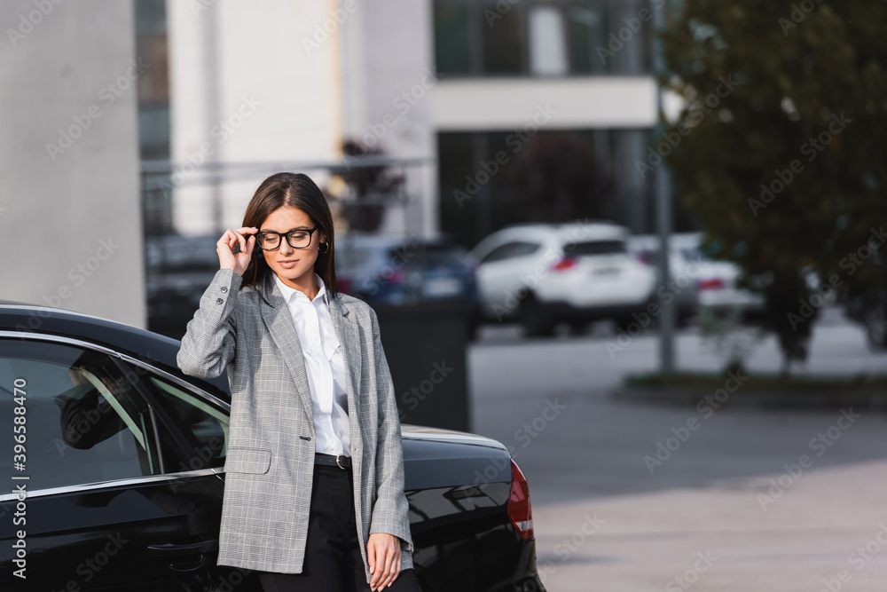Fototapeta premium businesswoman touching eyeglasses while standing near black car