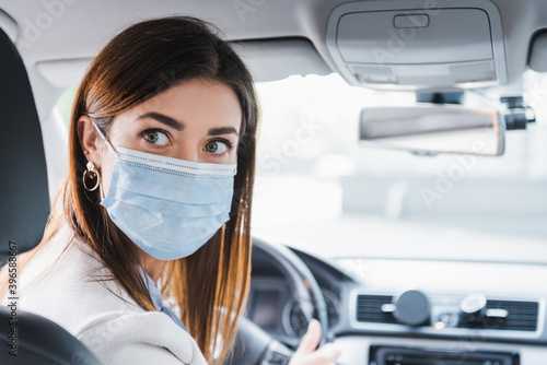  woman in medical mask looking back while driving car on blurred background