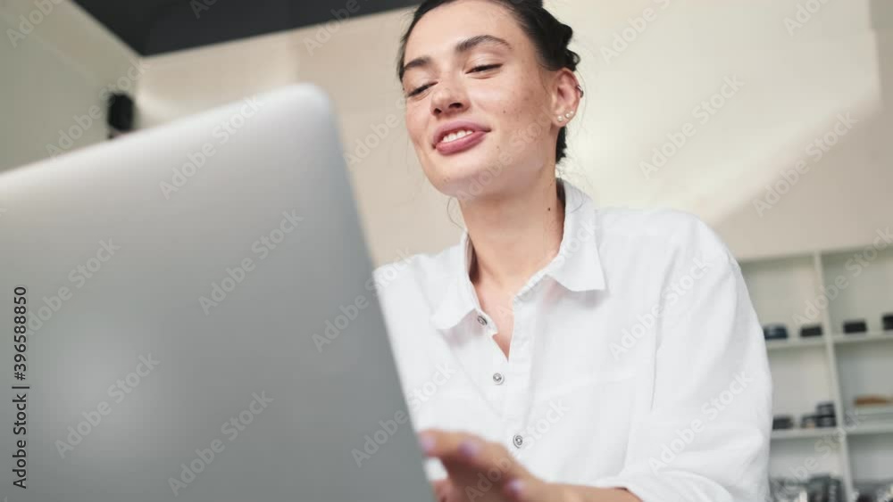 A smiling young woman is talking with someone using her laptop computer and video connection while sitting at the cafe indoors A smiling young woman is talking with someone using her laptop computer and video connection while sitting at the cafe indoors