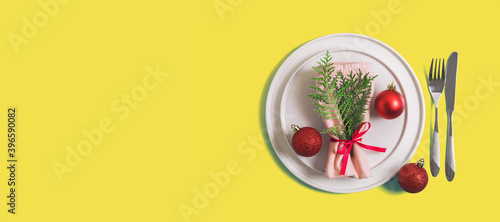 Website banner with served plate and cutlery for celebration of Christmas and New Year. On plate is napkin with Christmas tree branch, red balls. Flatlay on illuminating yellow background. Top view.