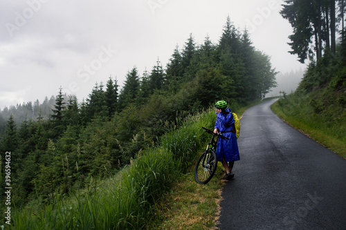 Young cycling woman standing on a road in raincoat have a rest in hill after riding a bike.