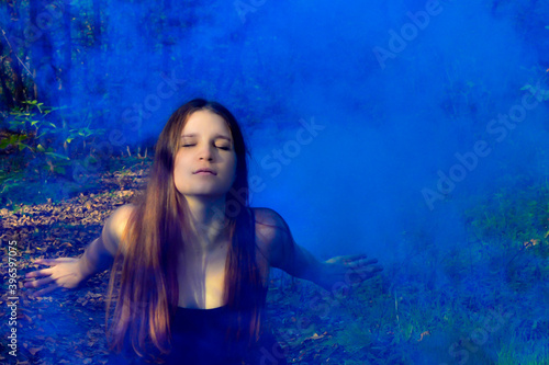 Red-haired woman sitting in a black long dress in a puff of blue smoke in the forest. Mysticism