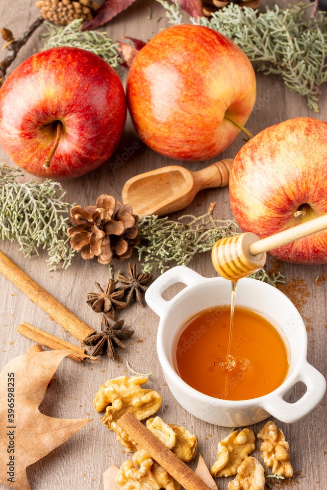 Top view of small white bowl with honey, red royal gala apples with cinnamon sticks, walnuts and autumn leaves, with selective focus, on wooden table, vertical