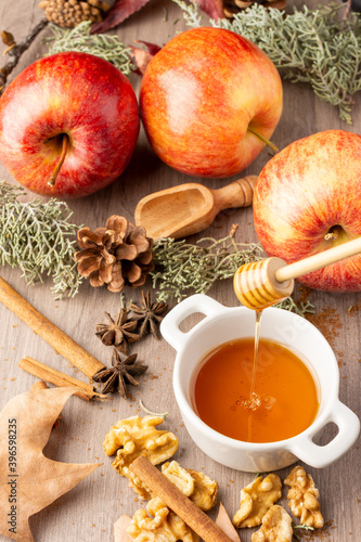 Top view of small white bowl with honey, red royal gala apples with cinnamon sticks, walnuts and autumn leaves, with selective focus, on wooden table, vertical