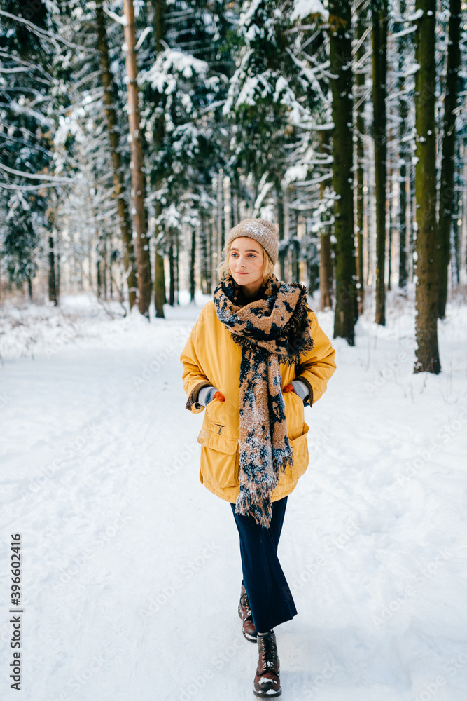 Fototapeta premium Young stylish hipster girl in yellow jacket with a warm scarf walking in the snow forest