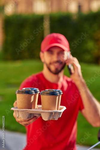 Young caucasian male courier talking by smartphone while delivering four coffee cups to custumer photo