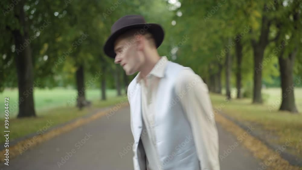 Camera moves up along handsome Caucasian man in elegant outfit strolling and dancing in summer park. Portrait of young confident guy enjoying sunny weekend outdoors, and leaving. Camera moves up along handsome Caucasian man in elegant outfit strolling and dancing in summer park. Portrait of young confident guy enjoying sunny weekend outdoors, and leaving.