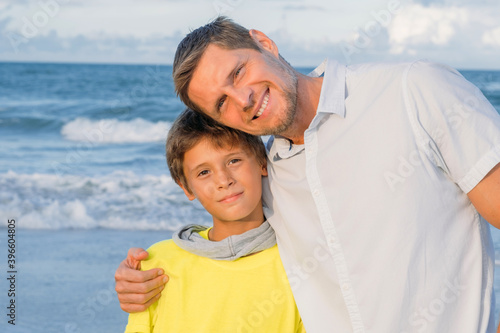 Father gently hugs his son and smiles happily into the camera against the background of the blue sea.