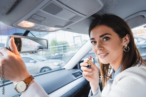  woman holding lipstick and looking at camera while adjusting rearview mirror in car on blurred foreground