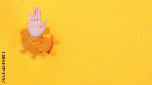 Bild auf Leinwand Woman hand arm showing stop gesture with palm isolated through torn yellow wall background studio