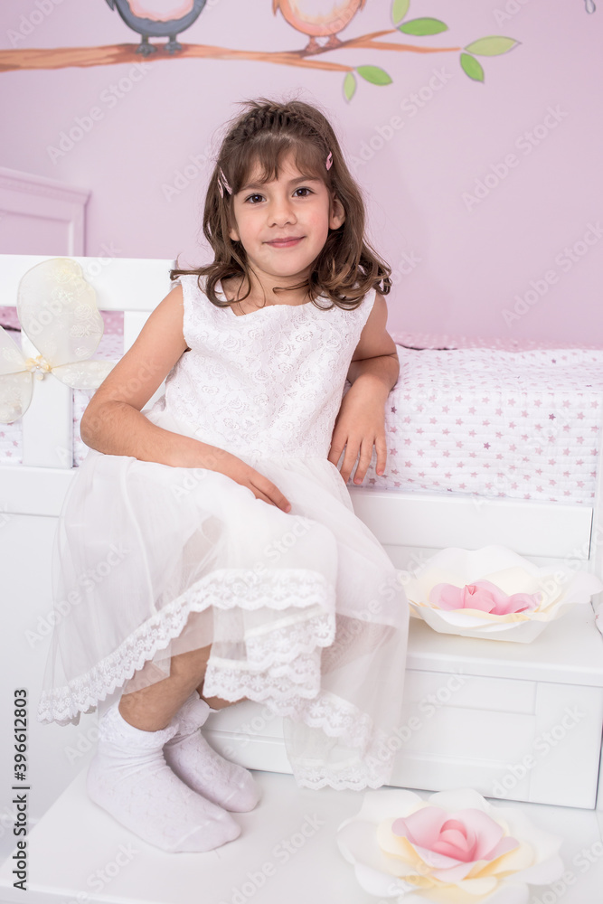 Little brunette girl sitting on her bed in white dress. Looking charming Little brunette girl sitting on her bed in white dress. Looking charming