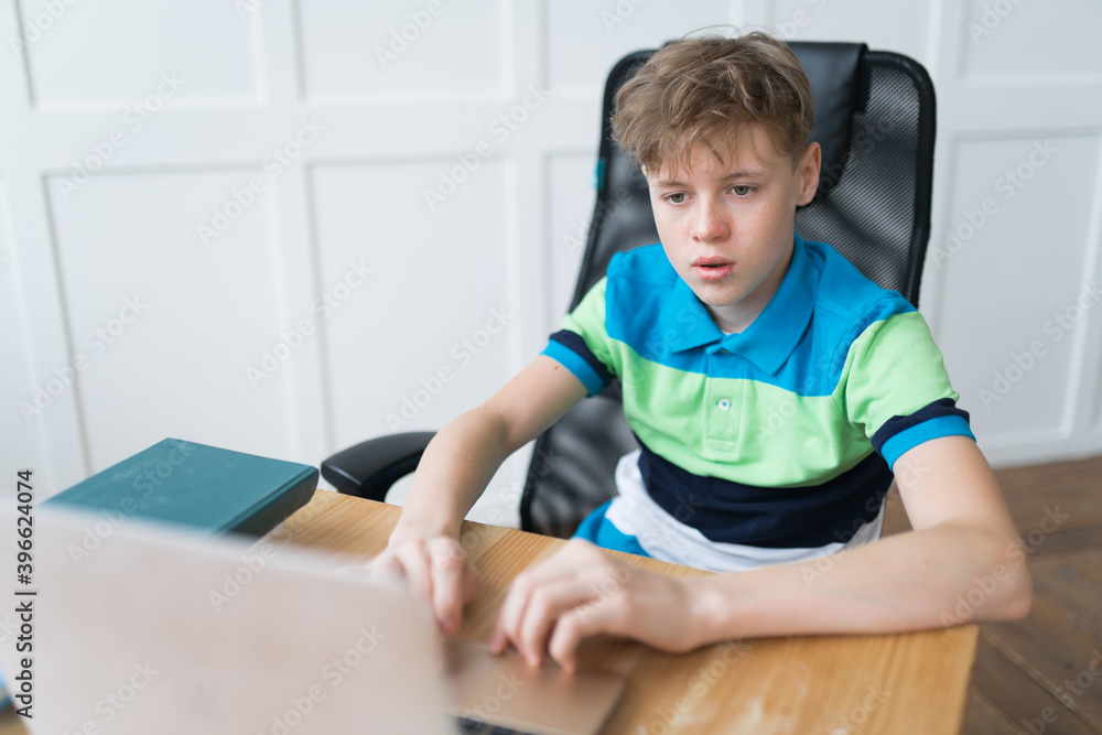 Caucasian schoolboy sitting at the desk and typing a text on his laptop, sending homework. He's focused, concentrated, confident Caucasian schoolboy sitting at the desk and typing a text on his laptop, sending homework. He's focused, concentrated, confident