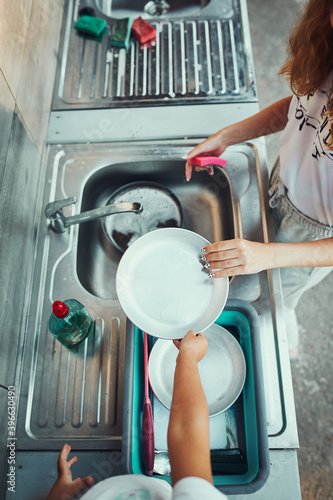 Fotografie Teenager girl washing up the dishes pots and plates with help her younger sister in the outdoor kitchen during vacations on camping