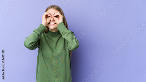 Young blonde woman isolated on purple background showing okay sign over eyes © Asier