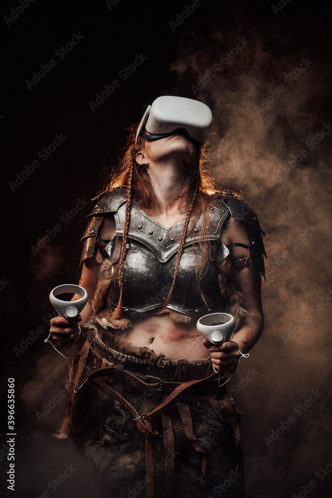 Fototapeta premium Barbaric scandinavian woman fighter in light armour with virtual reality headset and joysticks poses in dark smokey background.