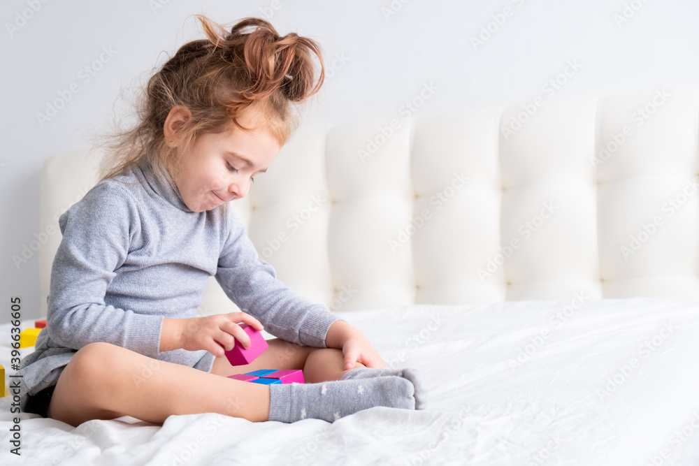 Little girl playing with colorful wooden cubes sitting on bed. Home activities for kids. Little girl playing with colorful wooden cubes sitting on bed. Home activities for kids.