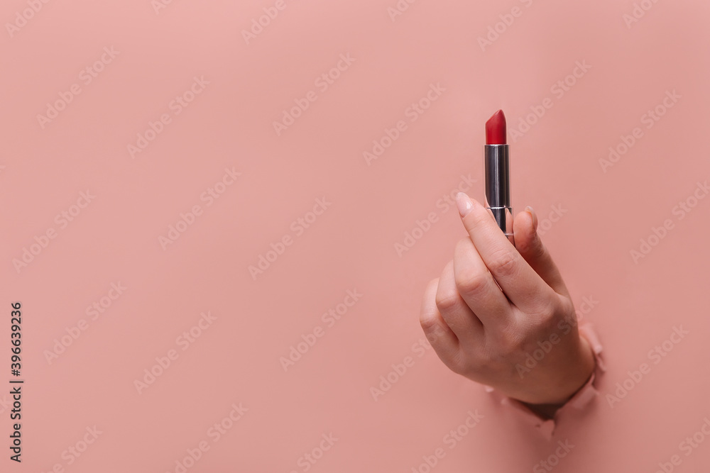 A female hand holds sexy red lipstick. Hole in torn paper trendy beige background. Make-up artist profession concept. Copy space for text banner. glamor accessory. Makeup accent A female hand holds sexy red lipstick. Hole in torn paper trendy beige background. Make-up artist profession concept. Copy space for text banner. glamor accessory. Makeup accent