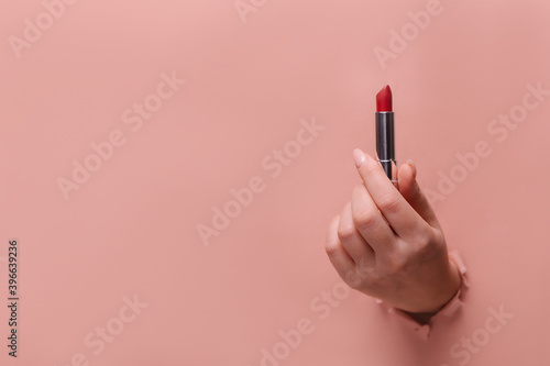 A female hand holds sexy red lipstick. Hole in torn paper trendy beige background. Make-up artist profession concept. Copy space for text banner. glamor accessory. Makeup accent
