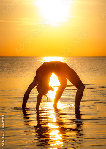 Fit girl demonstrates yoga asana Urdha Dhanurasana on the sea beach in the sunset light.