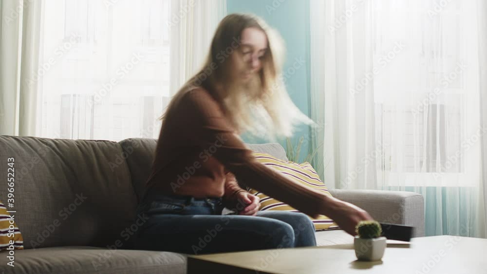 Young lady in blue jeans and brown sweater walks into room, sits down on sofa and picks up phone. Young lady in blue jeans and brown sweater walks into room, sits down on sofa and picks up phone.