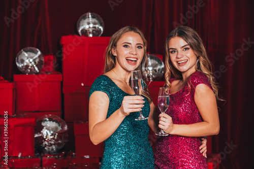 Two cheerful happy girls having a new year party, celebrating christmas. They drink champagne and open presents. They wear sexy dresses that curves their body in sexy way. Many red presents on back. photo