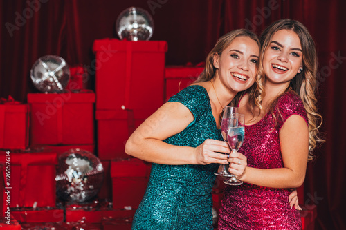 Two cheerful happy girls having a new year party, celebrating christmas. They drink champagne and open presents. They wear sexy dresses that curves their body in sexy way. Many red presents on back. photo