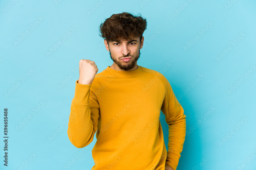 Young arab man on blue background showing fist to camera, aggressive facial expression. Young arab man on blue background showing fist to camera, aggressive facial expression.