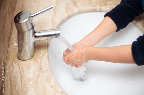 Young kid washing his hands and rinsing off soap from his hands under tap water during pandemia with rolled-up sleeves in a sink surrounded by brownish marble