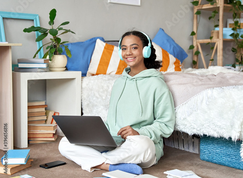 Tela Happy mixed race teen school girl distant college student wearing headphones virtual remote e learning using laptop in bedroom looking at camera