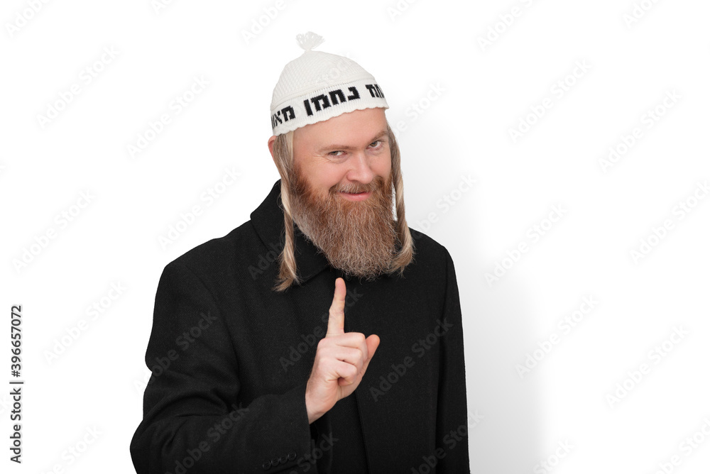 Smiling bearded jewish man with sidelocks in white kippah hinting do not try to deceive me. Charismatic sly jew dressed in black coat showing warning with finger on white background.