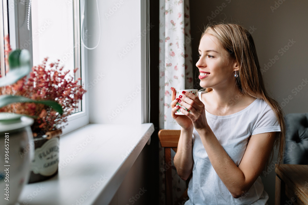 Adorable lovely romantic woman with long blond hair looking at window and smiling while drinking coffee and resting Adorable lovely romantic woman with long blond hair looking at window and smiling while drinking coffee and resting