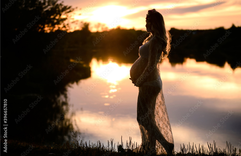 Pregnant woman in a gentle negligee near the river with a beautiful sunset. Beauty and tender motherhood.