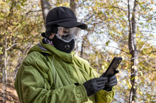 A woman wearing a respirator mask and protective glasses using a smartphone walking in the forest at COVID-19 period.