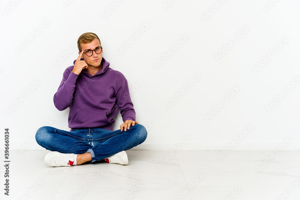 Young caucasian man sitting on the floor pointing temple with finger, thinking, focused on a task.