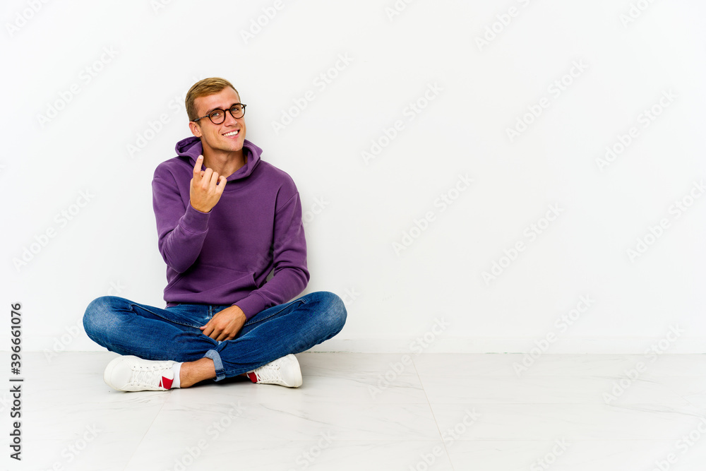 Young caucasian man sitting on the floor pointing with finger at you as if inviting come closer. Young caucasian man sitting on the floor pointing with finger at you as if inviting come closer.