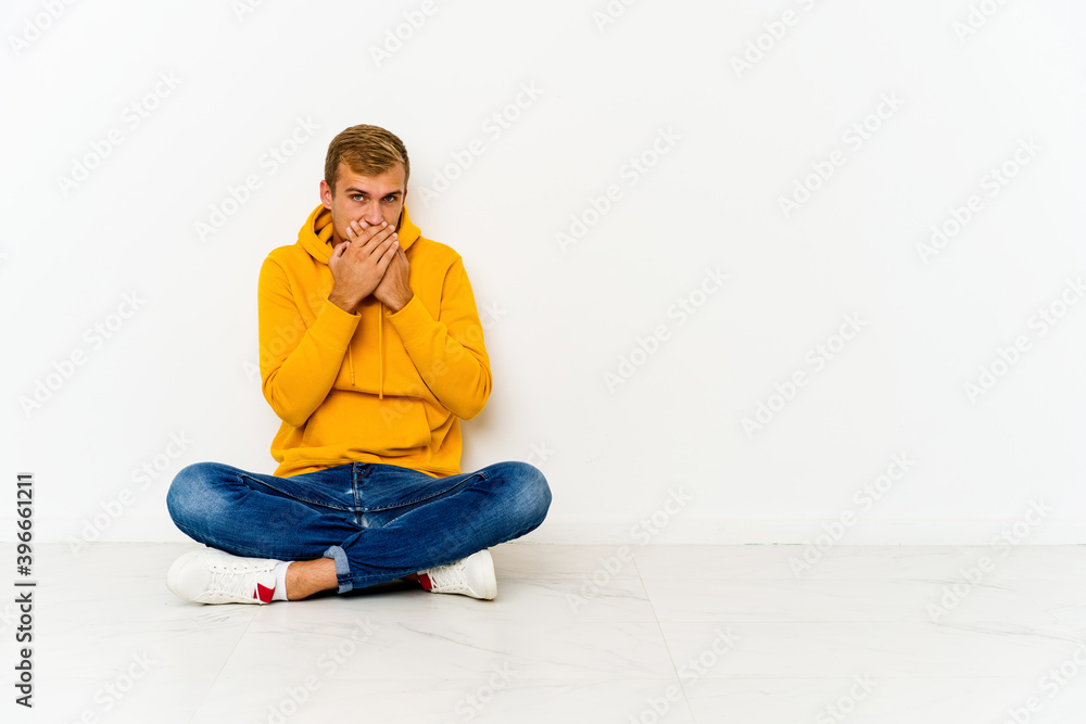 Young caucasian man sitting on the floor covering mouth with hands looking worried.