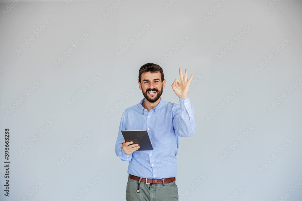 Smiling bearded young CEO standing in front of white background, holding tablet and showing okay sign while looking at camera. The business is going perfectly well. Smiling bearded young CEO standing in front of white background, holding tablet and showing okay sign while looking at camera. The business is going perfectly well.