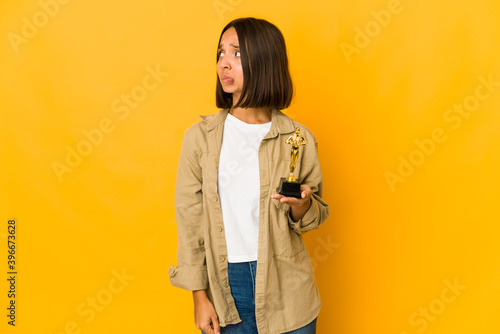 Young hispanic woman holding an award statuette confused, feels doubtful and unsure. © Asier