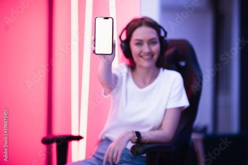 Smiling gamer girl wearing headset showing smart phone and pointing at blank screen with her finger.