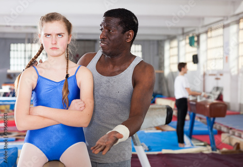 Male coach comforting sad teenage girl on gymnastic equipment at acrobatic hall