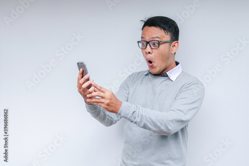 Wow face of Your Asian man shocked what he see in the smartphone on isolated grey background.