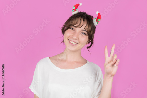 Portrait of a young woman with Christmas decorations in her hair, showing a peace sign with her fingers. Pink background. High quality photo