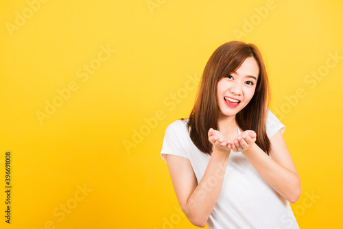 Asian happy portrait beautiful cute young woman teen standing wear t-shirt blowing kiss air something on hands looking to camera isolated, studio shot on yellow background with copy space
