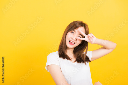 Asian happy portrait beautiful cute young woman teen smile standing wear t-shirt showing finger making v-sign symbol near eye looking to camera isolated, studio shot yellow background with copy space