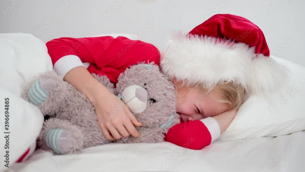 Smiling girl in Santa hat and Christmas pajamas lying in bed with teddy bear at home. New Year and Holidays. Christmas happiness morning Smiling girl in Santa hat and Christmas pajamas lying in bed with teddy bear at home. New Year and Holidays. Christmas happiness morning