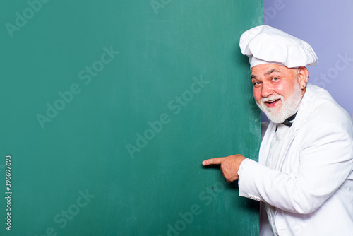 Senior chief cook. Portrait of chief cook with hand sign on blackboard background. Excited man chef cook wearing uniform showing copy space text. © Volodymyr