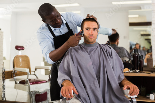 Male client getting trendy haircut at barber shop from African-American hairstylist using machine