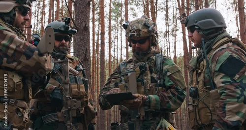A group of middle-aged bearded soldiers in uniforms and tactical vests discusses the action plan and prepares for action in a dense forest. The commander gives instructions to the soldiers.