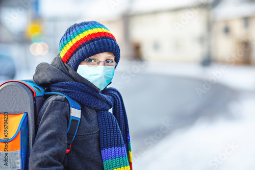 School kid boy with glasses wearing medical mask on the way to school. Child backpack satchel. Schoolkid on winter day with warm clothes. Lockdown and quarantine time during corona pandemic disease